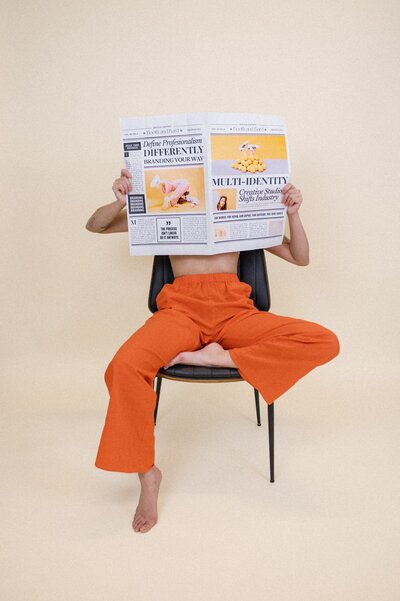 Woman wearing orange pants and no top sits in a black chair with a cream backdrop holding up a branded newspaper