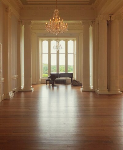 A large grand piano sits in a luxurious spacious empty room underneath a glowing chandelier. It's sunset and light floods the colonnaded and windowed room.