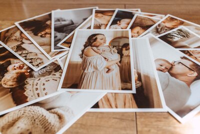 A collection of printed family photos is spread out on a wooden surface, featuring adults and babies in warm, neutral-toned clothing.