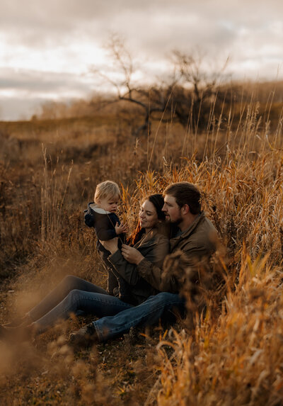 A family sits in a field of tall, dry grass. The woman holds a small child, and the man, seated beside them, looks on. Captured by a New England photographer, the warm and earthy setting with bare trees under an overcast sky suggests autumn's embrace.