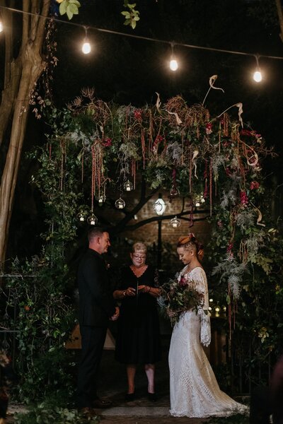 A couple standing under a lush, green floral arch with hanging lights during an evening wedding ceremony, accompanied by a woman officiating.