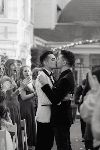 A newlywed couple shares their first kiss while guests cheer, captured in a candid and emotional cinematic wedding photo.