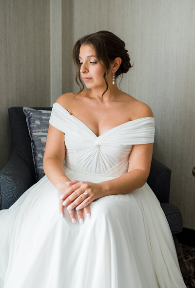Bride wearing off the shoulder wedding dress sits in chair while admiring her ring.