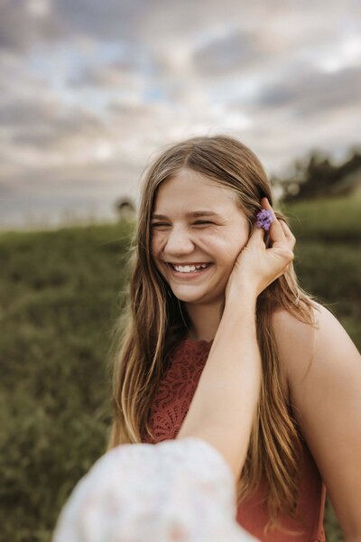 A girl with long hair smiles as another person places a purple flower behind her ear outdoors, captured beautifully by a Macon GA Photographer in a grassy field under a cloudy sky.