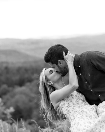 A couple twirls gracefully on a grassy hill, captured by an elopement photography New England expert. The woman in a flowing dress and the man in suspenders overlook scenic valleys with trees and houses, against rolling hills under a cloudy sky. The timeless black and white image captures their love.