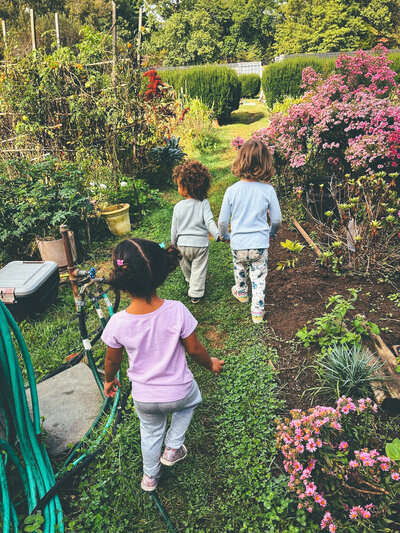 Three young children walking through a lush garden with blooming flowers, part of the Nests program for ages 0-4.