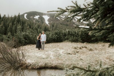 A couple in the distance with a mountain range in the background and a pond in front of the couple with the woman standing slightly behind the man with her head on his shoulder