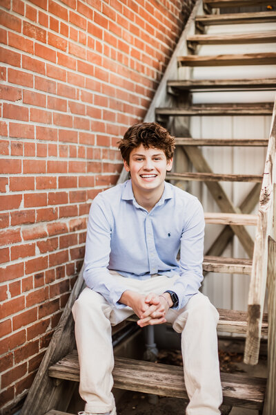 A person smiling while sitting on outdoor wooden stairs beside a brick wall.