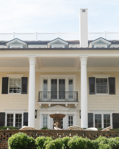 Photo of a private residence with large white columns and black shutters on the windows surrounded by a water fountain with lush greenery.