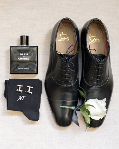 A pair of black dress shoes with a white rose on top, beside a bottle of Bleu de Chanel perfume, a pair of cufflinks, and black socks with initials AT embroidered on them, all arranged on a light-colored surface.