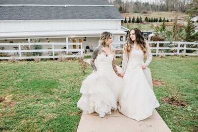 Two beautiful brides hold hands as they walk together on a foggy wedding day, their elegant gowns flowing, celebrating their love in a dreamy, romantic setting.