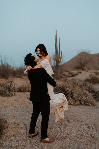 A couple in wedding attire are in the desert of Phoenix, Arizona for their elopement. The groom has lifted the bride of the ground as they look at each other and smile.