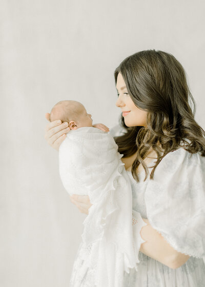 In studio newborn photo of a mother standing up and holding her newborn baby boy close and smiles.