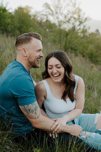 Engagement couple sitting in the grass during the summer laughing on the Blue Ridge Parkway