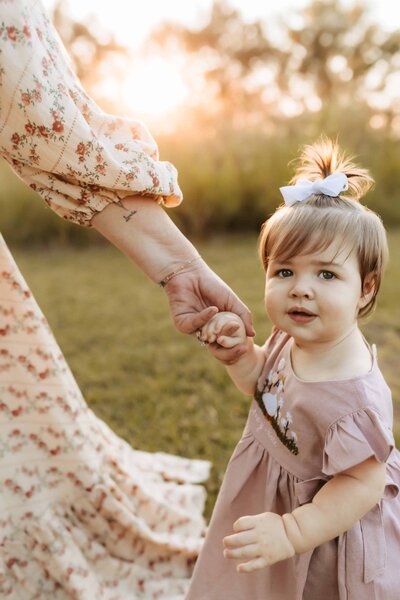A toddler in a pink dress holds an adult's hand outdoors at sunset, with blurred greenery in the background, beautifully framed by a Macon family photographer.