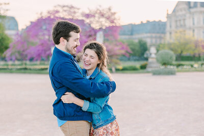 Couple laughing together as the man holds the woman during a surprise proposal at the Louvre and Tuileries Garden in Paris.