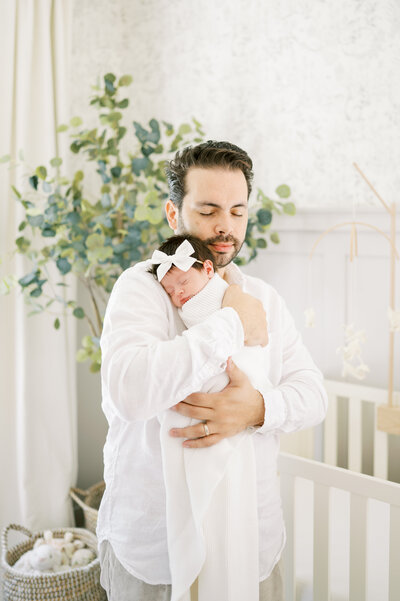 Dad snuggles newborn baby girl wrapped in white blanket during in-home photo session