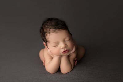 Baby boy is sitting in froggy pose for a New Jersey newborn photos. His hands are under his chin and his face is slightly angled away from the camera. He is sleeping peacefully. Captured by best NJ newborn photographer Katie Marshall.