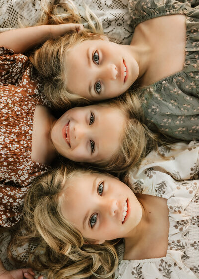 Three blonde young sisters laying head to head with toothy grins. 