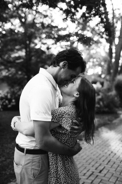 A couple embraces and smiles at each other on a stone path, surrounded by trees and greenery. Captured in classic black and white, this moment of joy is a testament to the timeless artistry of family film photography by Toronto family photographer, Jennifer van Son.