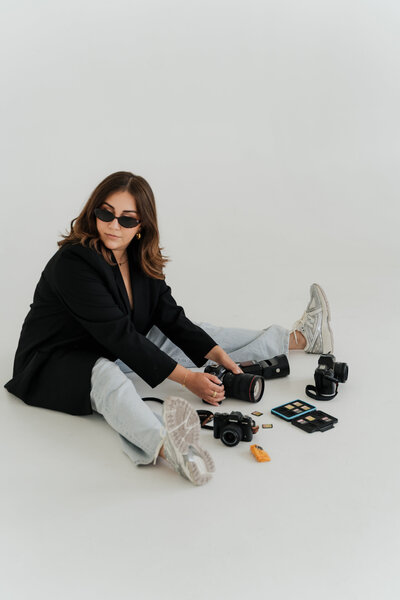 Woman sits on the floor of a white studio surrounded by film cameras, rolls of film, and a light meter, wearing sunglasses and sneakers.