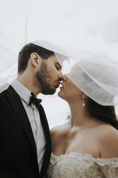 A bride and groom share a gentle kiss beneath a flowing veil, the groom in a black tuxedo and the bride wearing drop earrings and a strapless gown.
