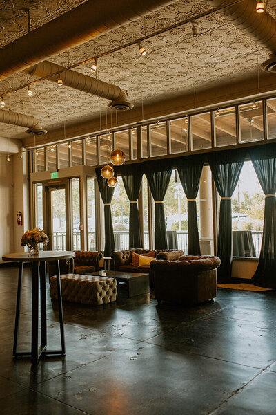A sunlit lounge area at SoHo63, a Chandler wedding and event venue, featuring tufted seating, dark drapes, warm lighting, and modern industrial touches—perfect for cocktail hour or intimate indoor gatherings.