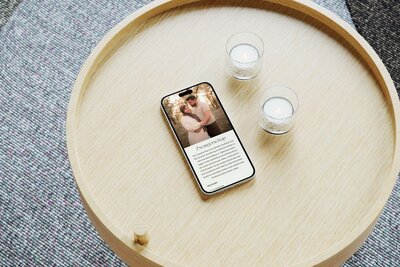 Smartphone lying on a round light-wood tray table beside two glass tealight candles, screen displaying a maternity photography page with a couple embracing in a sunlit forest.