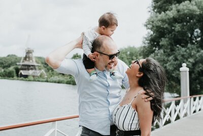 Family on lakeside pier in autumn – Smiling parents sit with their two young children and a fluffy dog on a wooden pier beside a calm lake and thatched-roof house, surrounded by golden fall foliage.