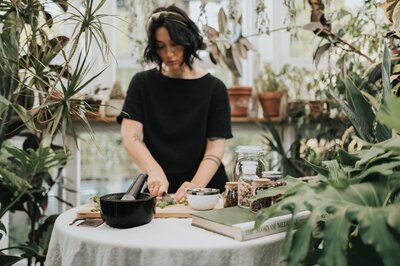 Woman stands in lush, greenhouse wearing a headband and black t-shirt while chopping herbs on cutting board atop a table with linen tablecloth.