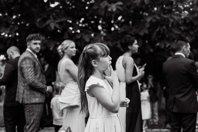 Stunning reportage style black and white wedding photo at same level side on of the flowergirl holding the confetti to her chin eagerly awaiting the bride and groom