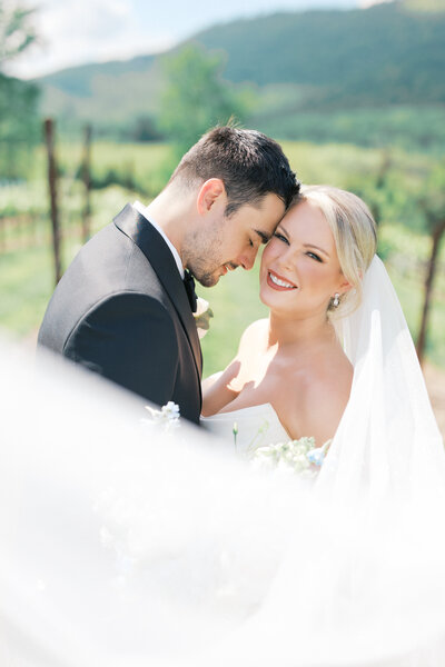 Groom rests into blond haired Bride's temple and she smiles and veil flows across 
