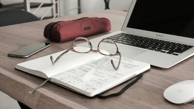 Workspace setup with an open notebook filled with handwritten notes, as well as eyeglasses, pen, laptop, pencil case, smartphone, and mouse on a wooden desk at Kidwell Coaching