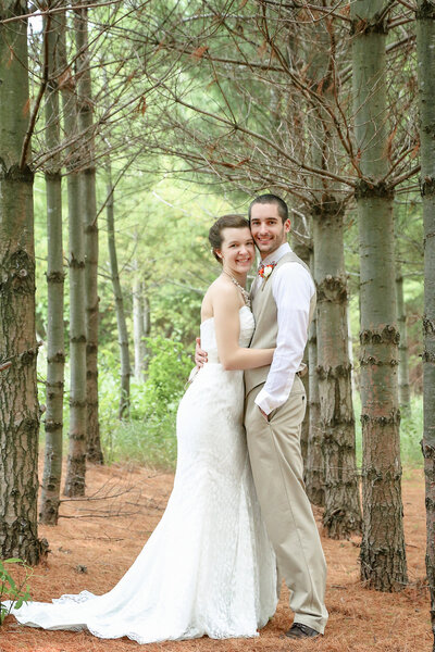 A white couple wearing wedding attire standing in a pine forest looking at the camera