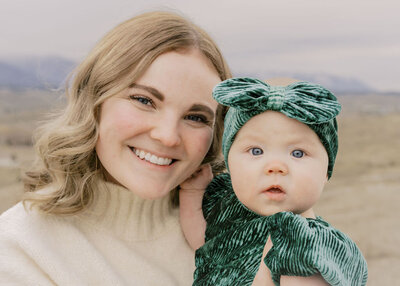Smiling mother holding her baby dressed in green with a matching bow, captured by Joseph Mayden during a natural light family photography session in a mountain setting.
