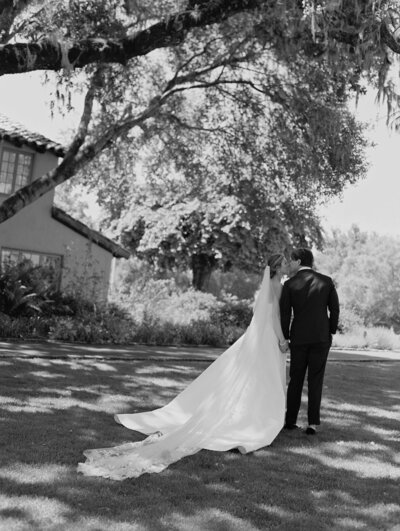 A bride walking on grass with lace top of dress and long train of gown behind her. 
