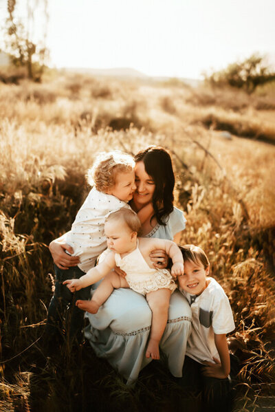 Mom sits in a field at sunset with her three babies in her lap.
