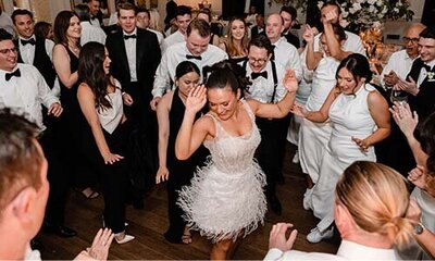 Bride in feathered white mini dress leading an energetic dance floor surrounded by wedding guests