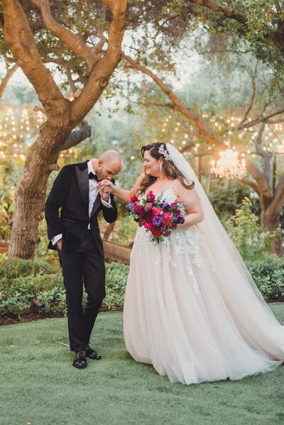 groom romantically kisses brides hand in garden with chandeliers