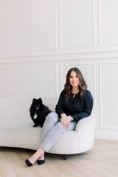 Brunette woman sitting on white couch next to black Pomeranian with a tie on.