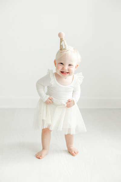 One year old baby girl smiles joyfully in white dress and birthday hat during birthday portrait session