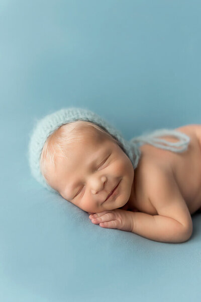 Baby boy in bonnet sleeps on stomach on a teal backdrop for his newborn photos.