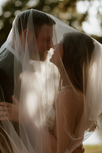 A bride and groom share an intimate moment, smiling under a softly lit veil. The scene conveys romance and joy, set against a blurred natural backdrop.