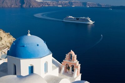 An ocean crusie ship sails on the blue water of the mediterranean in teh background while the viewer looks out over blue and white stucco buildings on the edge of a Greek island.