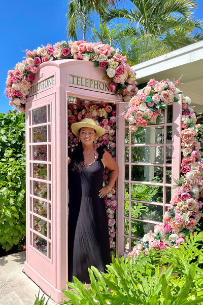 A woman in a black dress posing in a pink floral-covered phone booth, capturing the essence of stylish travel and destination weddings.