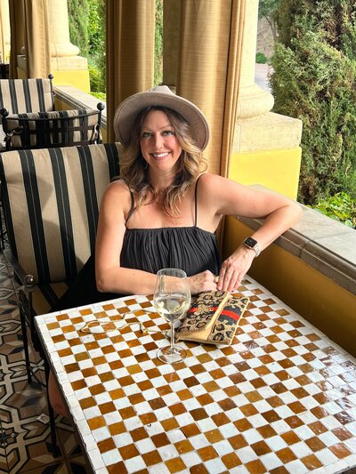 Anne sitting at a table with a drink near a window