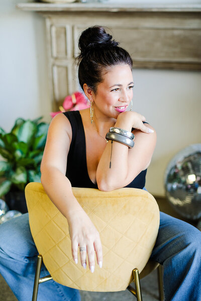 woman with high bun and black tank top sits backwards on plush yellow chair. her hand is resting on her chin.