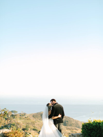 Bride and groom embrace on a cliff overlooking the ocean under a clear blue sky, creating a serene and romantic atmosphere.