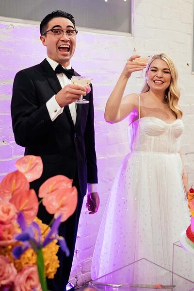 Smiling bride and groom raising cocktails for a toast at their wedding reception, surrounded by colourful florals and soft lighting.