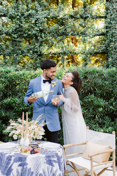 Groom pours champagne while bride clutches his arm.